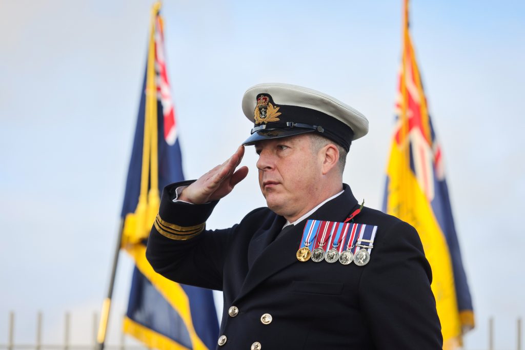 Naval Commander in full uniform saluting with standards in the background